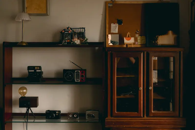 A wooden cabinet and display shelf housing vintage film cameras, radios, and optical equipment in a room setting