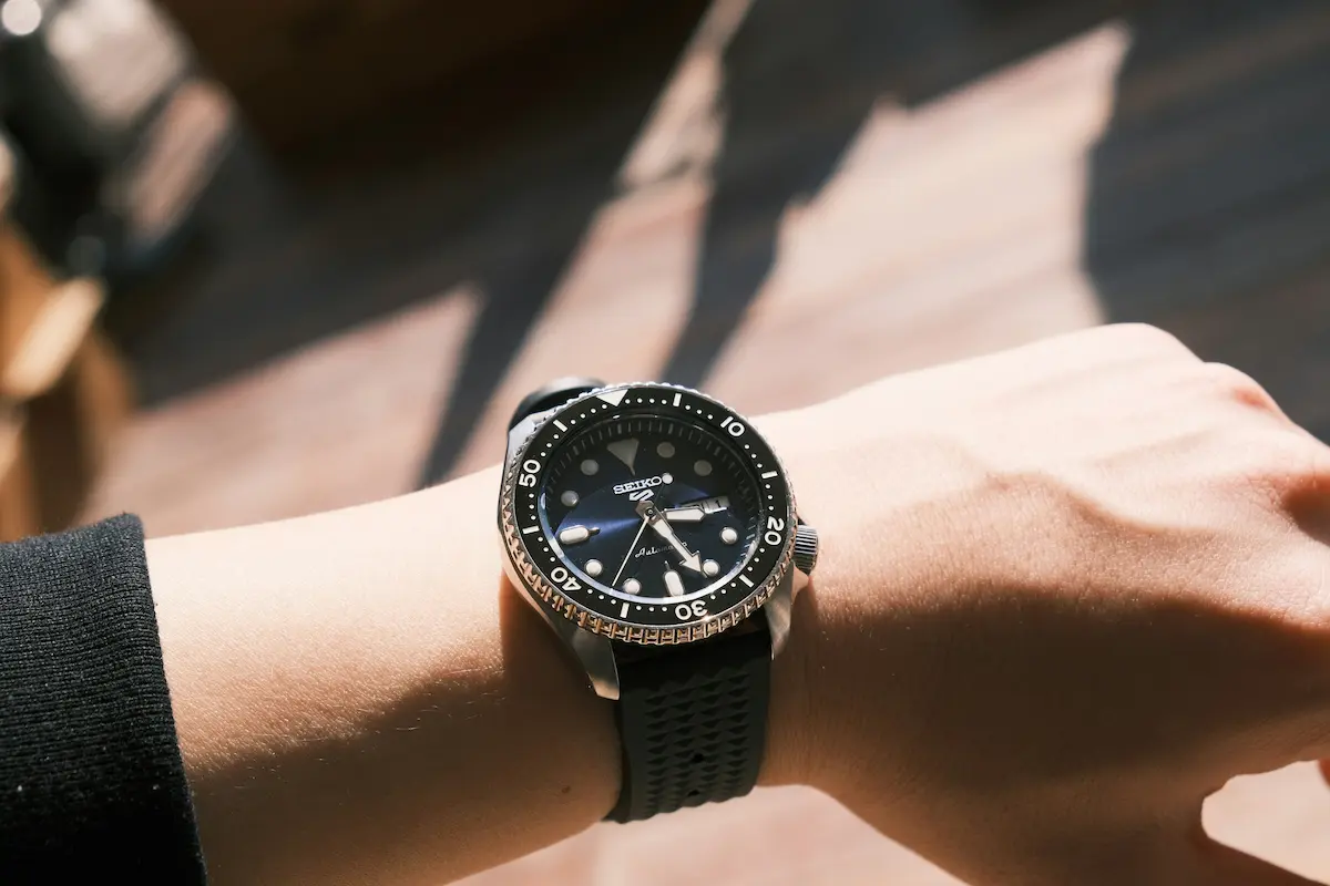 A close-up of a person wearing a black Seiko 5 Sports automatic watch with a textured rubber strap under natural sunlight