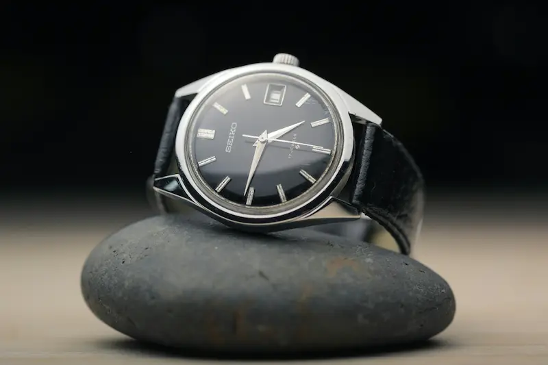 A classic vintage Seiko 17 Jewels manual-wind watch with a black dial and leather strap resting on a smooth black stone