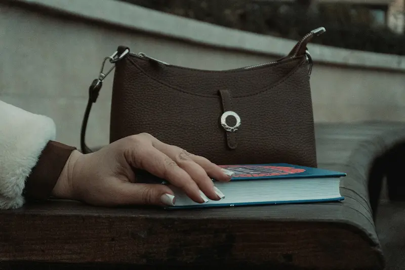 A brown leather designer handbag sitting on an outdoor wooden bench next to a book, representing everyday use and environmental exposure