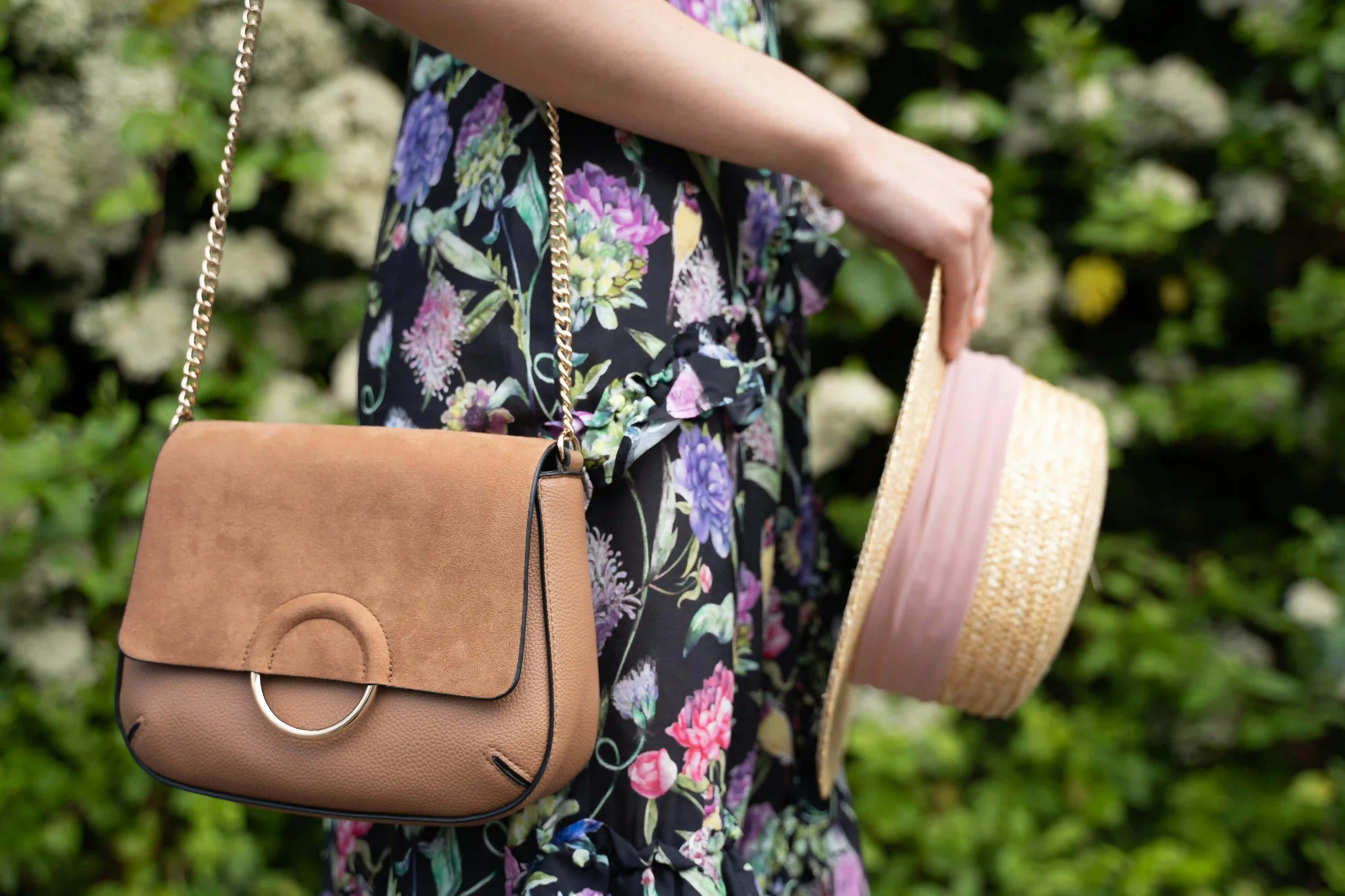 A woman wearing a floral dress styling a tan designer shoulder bag with a prominent suede leather flap and a gold chain strap
