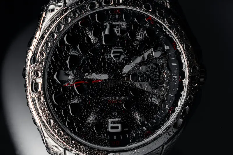 A macro close-up of a black Casio Edifice watch covered in heavy water droplets, displaying the Water Resist 100M rating on the dial