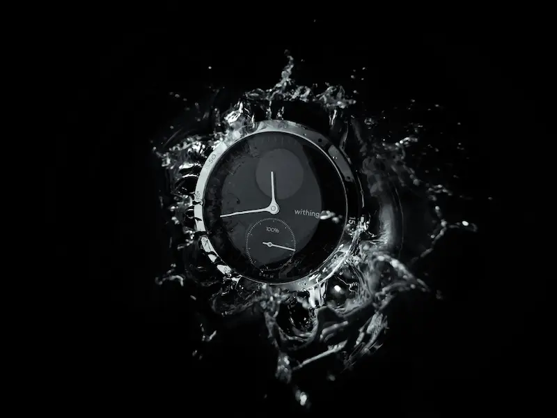 A black Withings hybrid smartwatch facing dynamic water splashes against a solid black background
