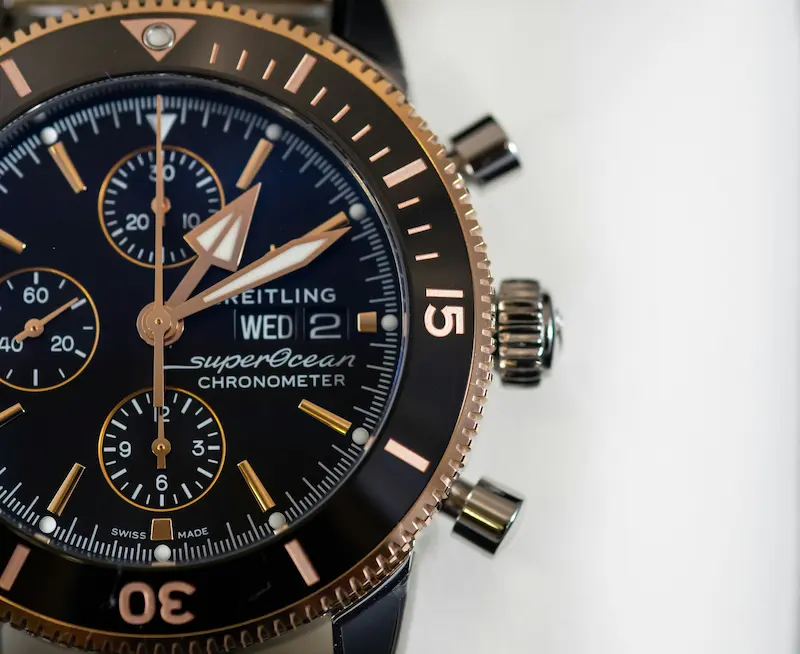 A macro close-up of a black and rose gold Breitling Superocean Chronometer watch dial, showcasing the chronograph sub-dials, day-date window, and crown buttons