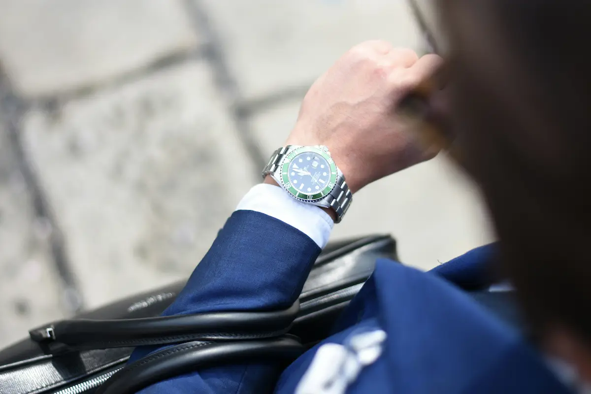 An over-the-shoulder view of a businessman in a blue suit checking a stainless steel Rolex Submariner watch with a green bezel on his wrist