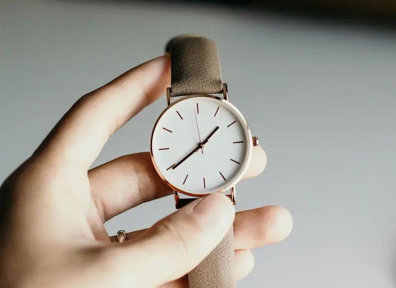 A close-up of a person's fingers holding and carefully inspecting a minimalist rose gold watch with a white dial and brown leather strap