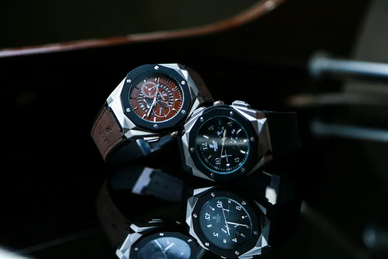 Two luxury Hublot watches with angular stainless steel cases sitting on a glossy black reflective surface