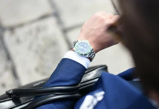 An over-the-shoulder view of a businessman in a blue suit checking a stainless steel Rolex Submariner watch with a green bezel on his wrist