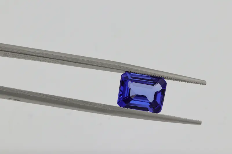 A close-up photograph of a loose, emerald-cut blue sapphire gemstone being held carefully by silver metal tweezers