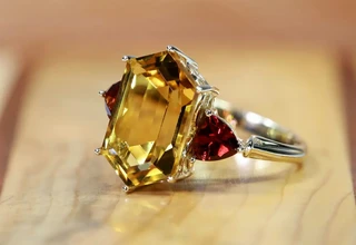 A macro close-up of a gold ring featuring a large emerald-cut yellow gemstone secured by metal prongs, flanked by two trillion-cut red gemstones