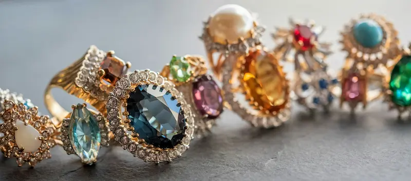 A row of various vintage gold and silver rings featuring colorful gemstones like blue topaz, amber, emerald, and pearls surrounded by diamond halos