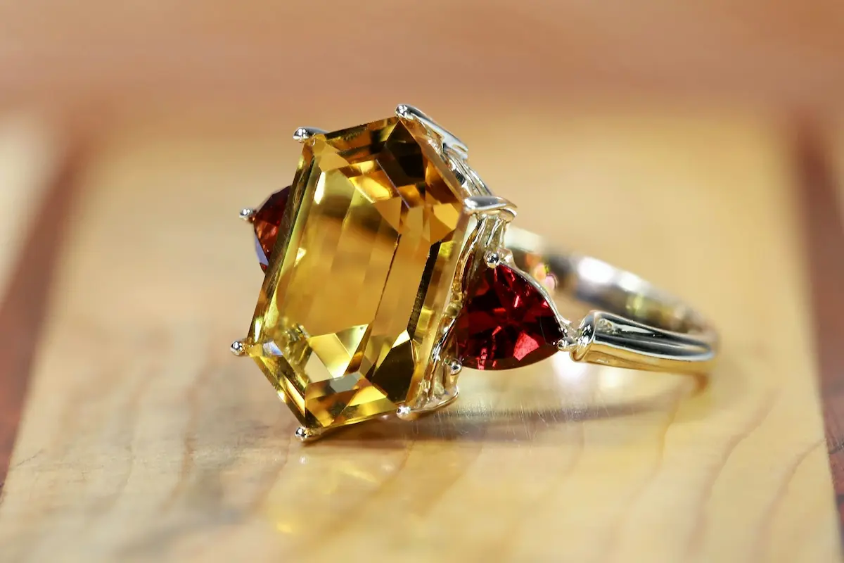 A macro close-up of a gold ring featuring a large emerald-cut yellow gemstone secured by metal prongs, flanked by two trillion-cut red gemstones