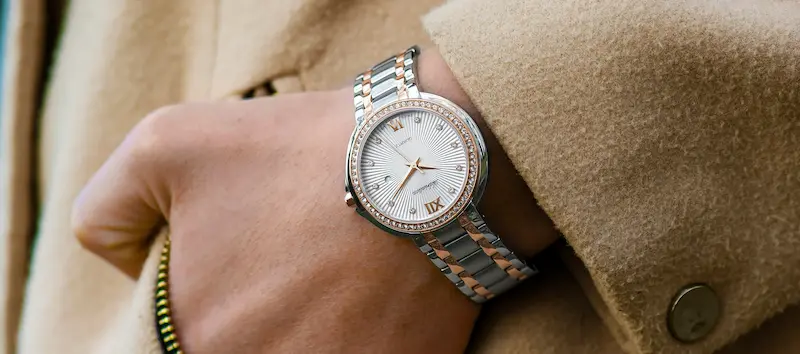 A close-up of a premium women's watch with a silver and rose gold link bracelet, diamond-studded bezel, and white textured dial, worn under a beige trench coat