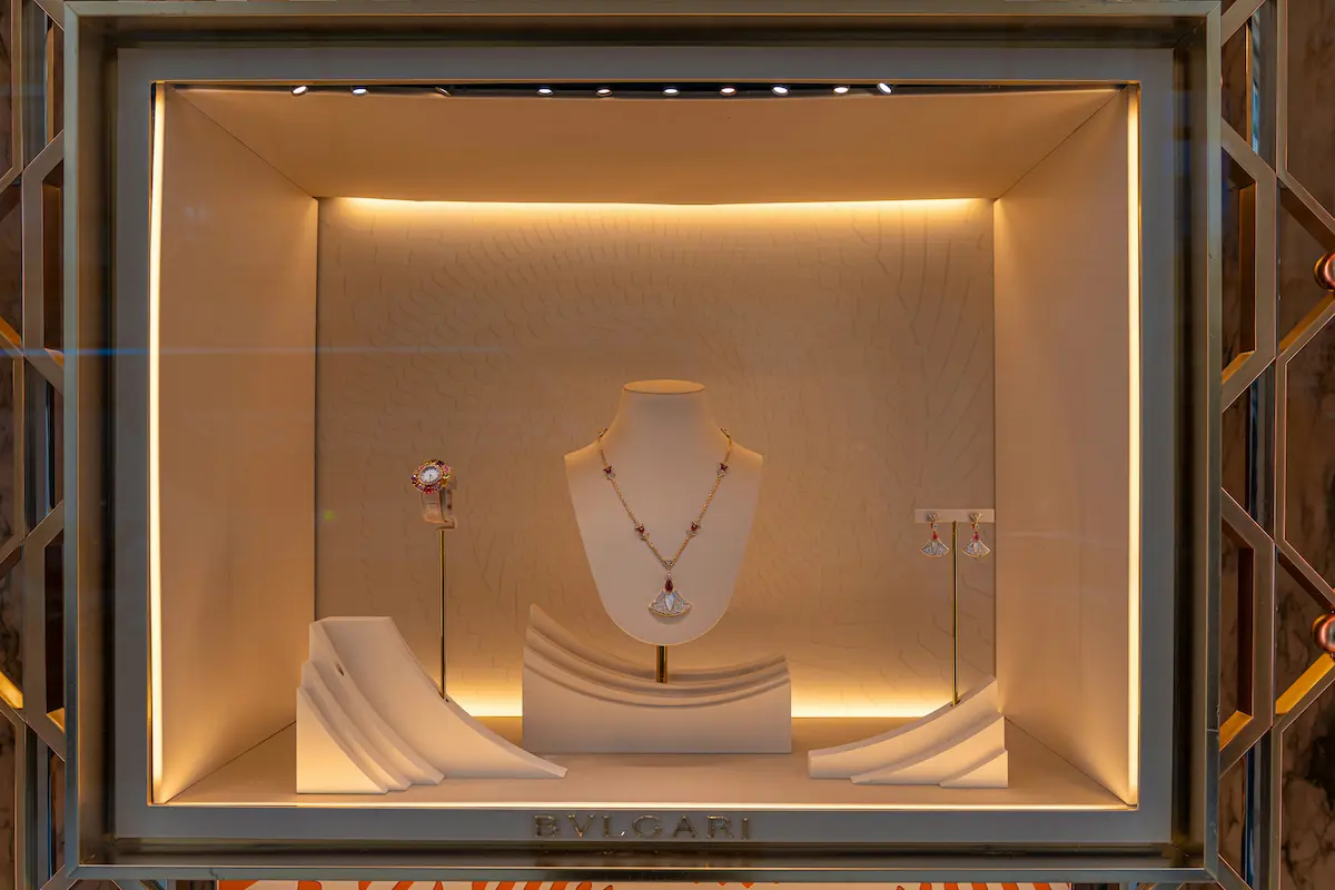 A luxury BVLGARI storefront display window showcasing a high-end jewelry collection including a diamond and ruby necklace, an ornate watch, and matching earrings on elegant white stands