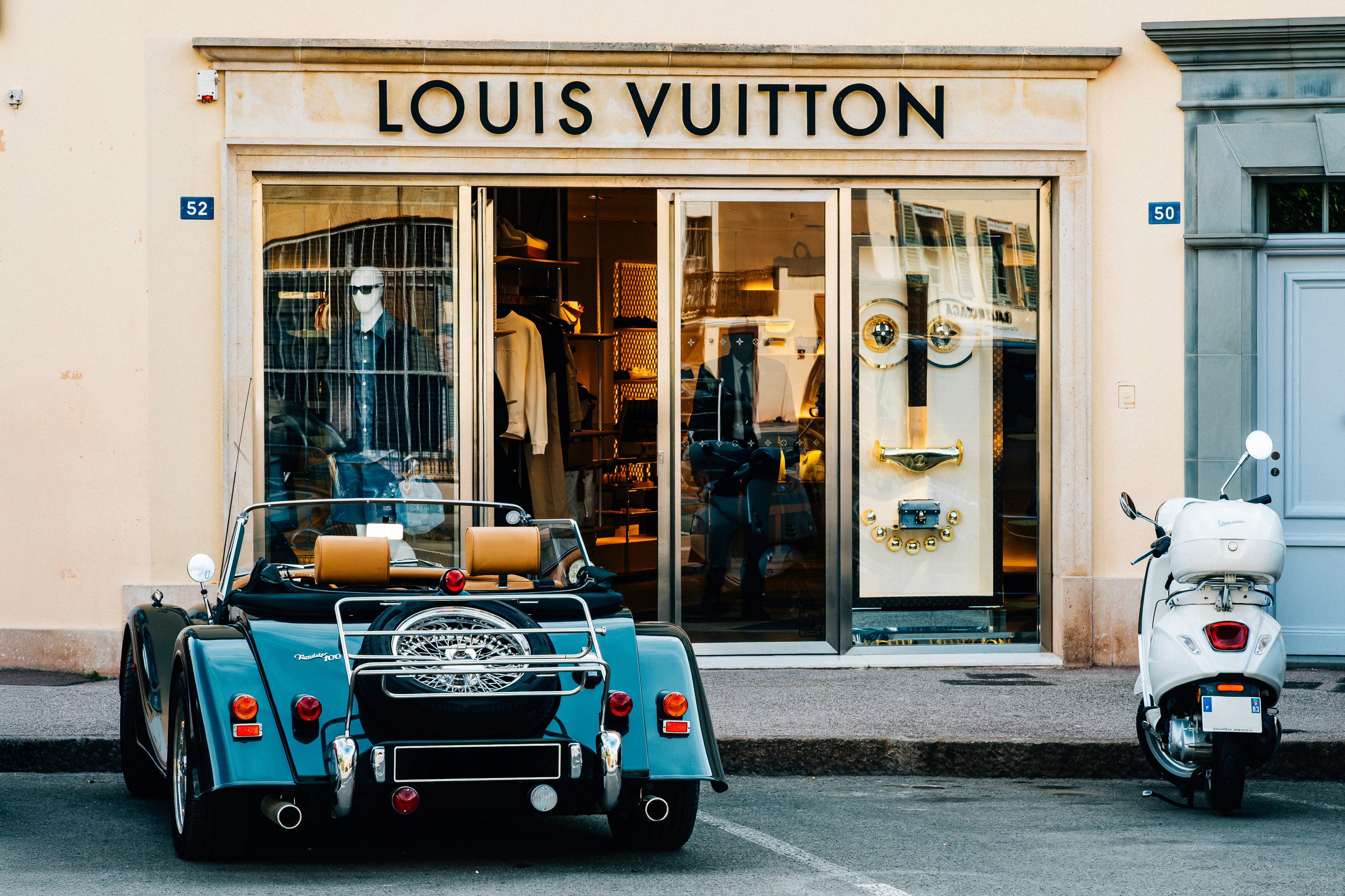 A classic dark green convertible car and a white Vespa scooter parked in front of a luxury Louis Vuitton storefront boutique