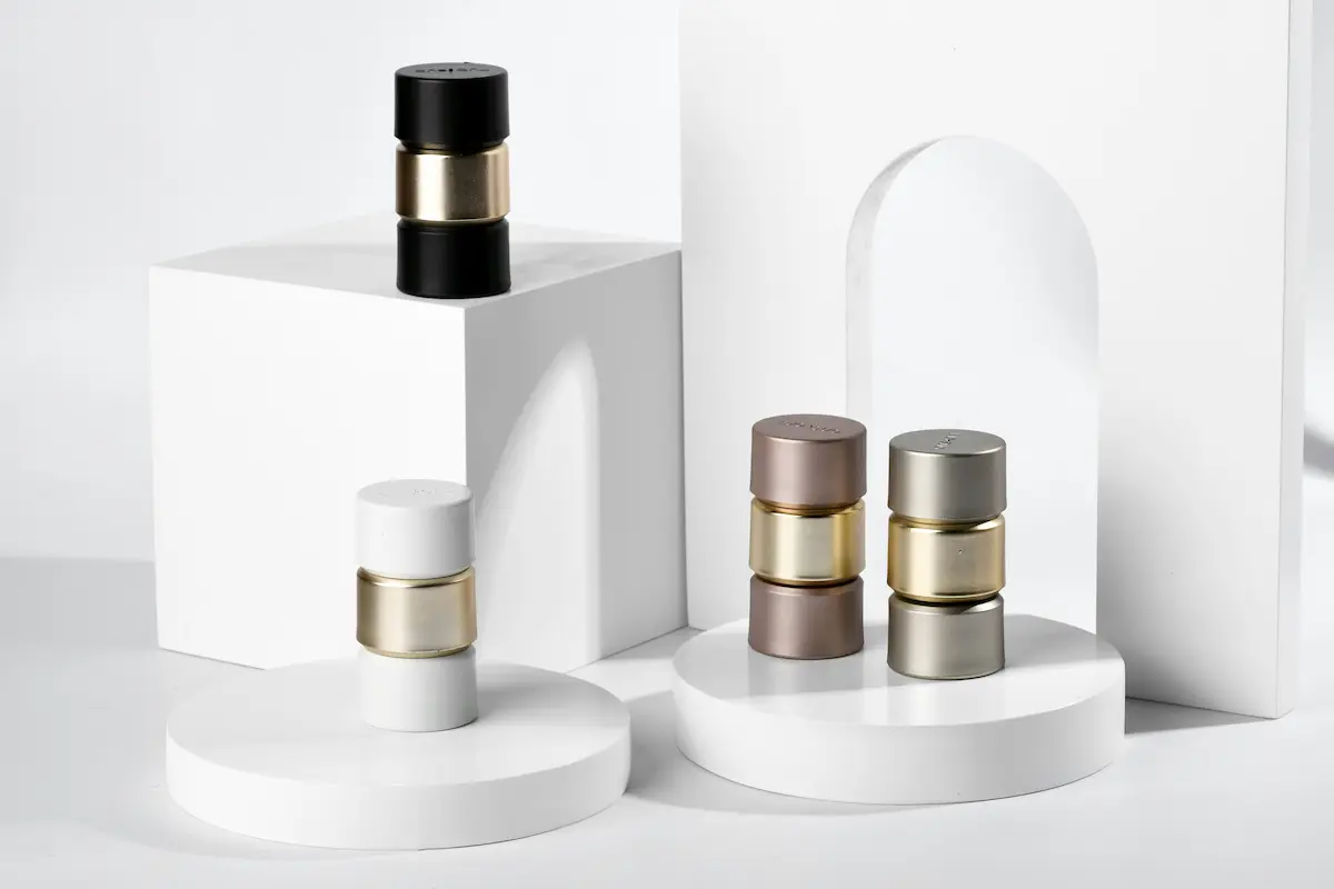 Four minimalist cylindrical metal perfume atomizers in black, white, mocha, and silver gray with gold center rings, arranged on white geometric product display risers