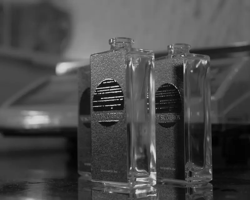 A black and white close-up of empty glass perfume bottles with texturized metallic labels featuring the word MUSKAMBROX, showing open necks without spray nozzles