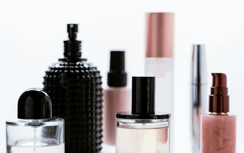 A collection of various perfume and cosmetic bottles, including clear glass perfume vials, a studded black bottle, and metallic spray atomizers against a white background