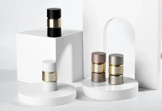 Four minimalist cylindrical metal perfume atomizers in black, white, mocha, and silver gray with gold center rings, arranged on white geometric product display risers