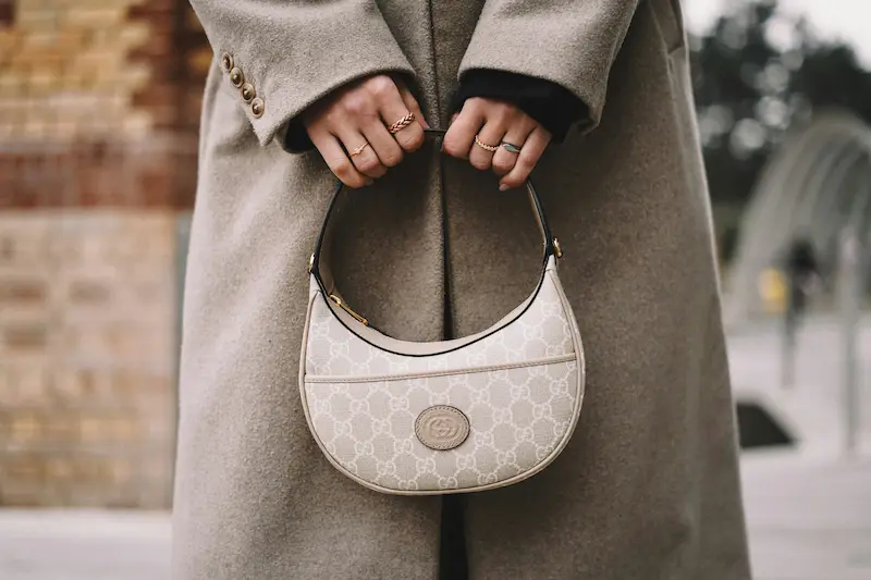 A close-up street style photograph of a woman wearing a beige wool coat, holding a cream Gucci GG Supreme crescent hobo bag with ivory leather trim and structural stitching details