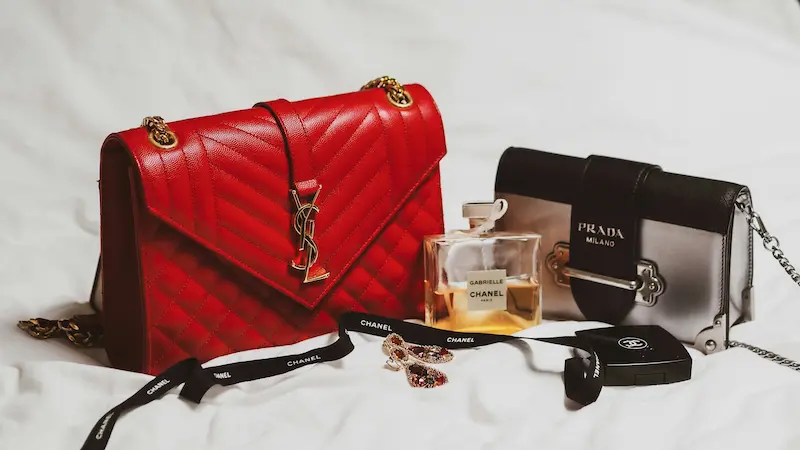 A luxury flat lay showcasing a red quilted YSL envelope shoulder bag with gold hardware next to a silver and black Prada Cahier chain bag and a Chanel perfume bottle on a white sheet