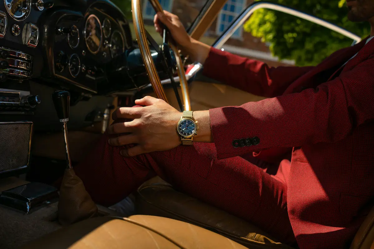 A close-up lifestyle shot of a man wearing a red suit inside a vintage car, showcasing a luxury gold case chronograph watch with a gold mesh bracelet on his wrist