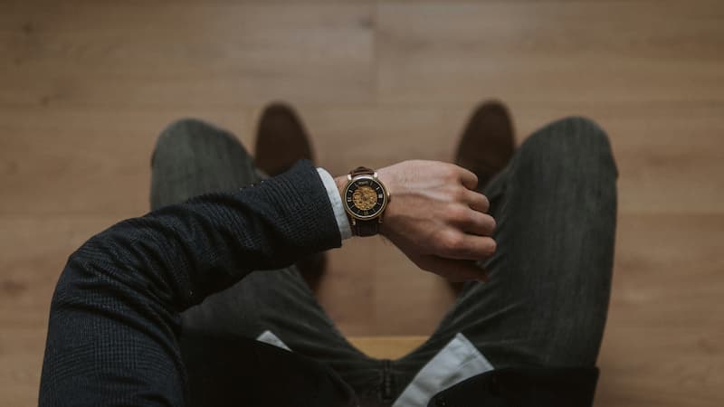 A top-down view of a man in a dark suit checking his luxury gold case skeleton watch with a brown leather strap, shot against a wooden floor background