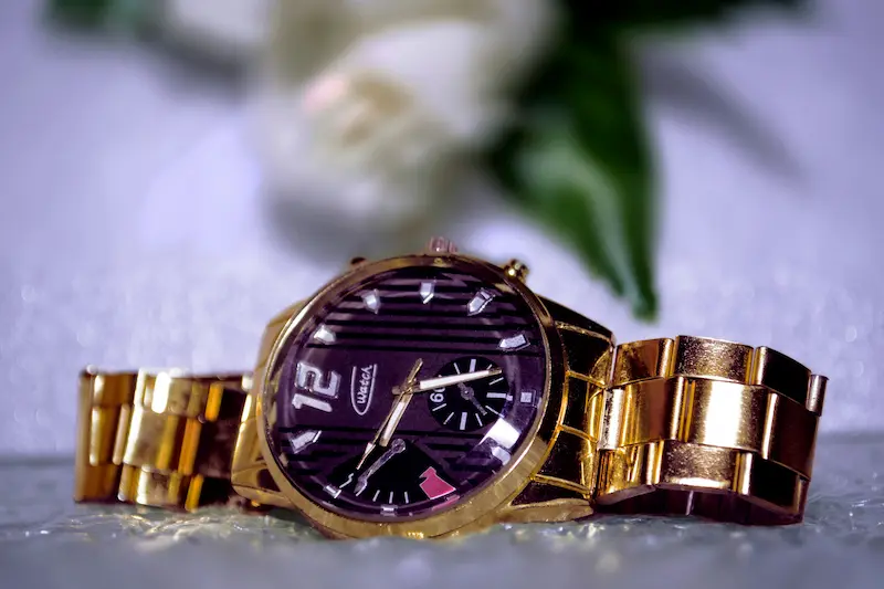A close-up studio shot of a polished gold watch with a textured black dial and a matching gold link bracelet resting on a reflective surface