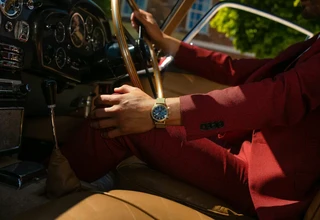 A close-up lifestyle shot of a man wearing a red suit inside a vintage car, showcasing a luxury gold case chronograph watch with a gold mesh bracelet on his wrist
