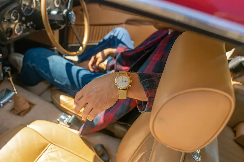 A close-up of a man's wrist wearing a vintage gold watch with a mesh bracelet, seated inside the tan leather interior of a classic car