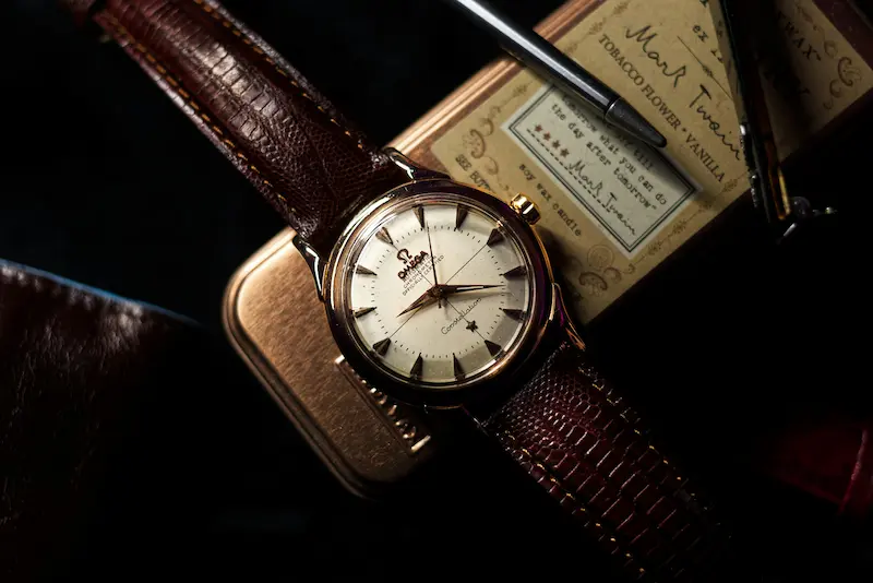 A vintage Omega Constellation automatic chronometer watch with a pie-pan dial and brown leather strap, resting on a brass box and a Mark Twain vintage candle tin