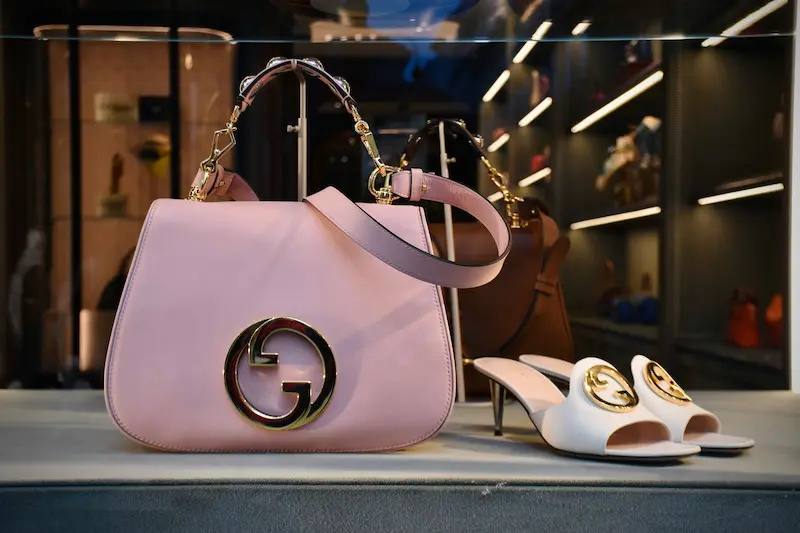 A pink leather designer luxury handbag with a prominent large round gold logo emblem and gold clasp attachments, displayed inside a high-end fashion boutique window next to white heels
