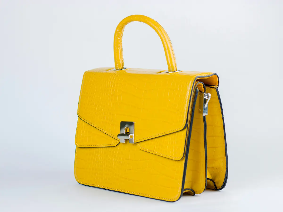 A vibrant yellow designer handbag with a faux crocodile leather texture, featuring a shiny silver clasp closure and metal hardware rings on a white background