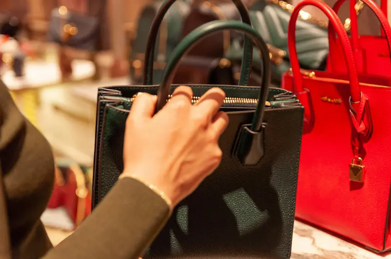 A close-up of a customer's hand touching the handle of a dark green leather luxury handbag with a prominent gold zipper, displayed next to a bright red bag in a boutique