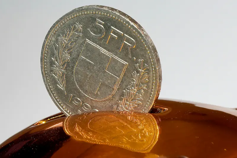 A silver 5 Swiss Franc coin being inserted into the slot of a glossy brown piggy bank, reflecting on its shiny surface against a plain light background