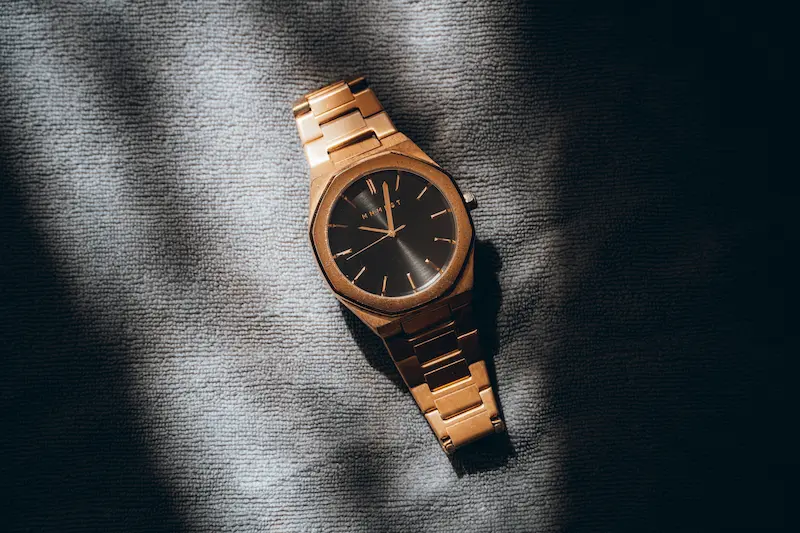 A gold-toned wristwatch featuring an octagonal gold case, dark black dial, and matching gold bracelet resting on a textured grey cloth surface with dramatic lighting