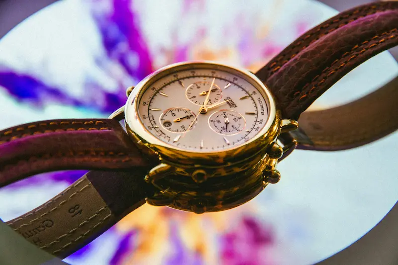 A polished gold-toned Citizen chronograph watch with a white dial and brown leather strap resting on a reflective mirror surface