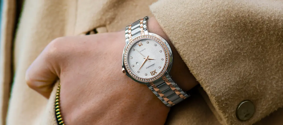 Close-up of a person wearing a two-tone silver and rose gold wrist watch with a crystal-embellished fixed bezel against a beige coat