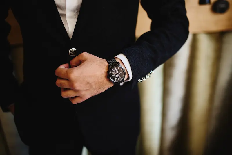 Close-up of a man in a black tailored suit adjusting his jacket, wearing a sleek all-black luxury chronograph watch with a mesh strap.