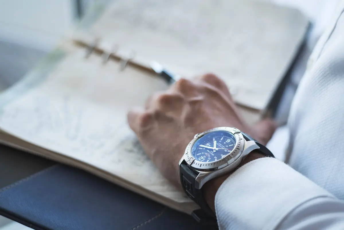 Close-up of a man's wrist wearing a stainless steel luxury chronograph watch with a black strap while holding an open planner.