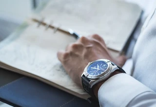 Close-up of a man's wrist wearing a stainless steel luxury chronograph watch with a black strap while holding an open planner.