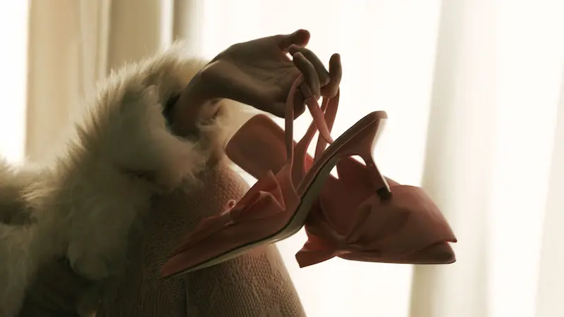 A hand wearing a soft fur cuff holding elegant pale pink slingback heels with subtle bow embellishments against soft lighting.