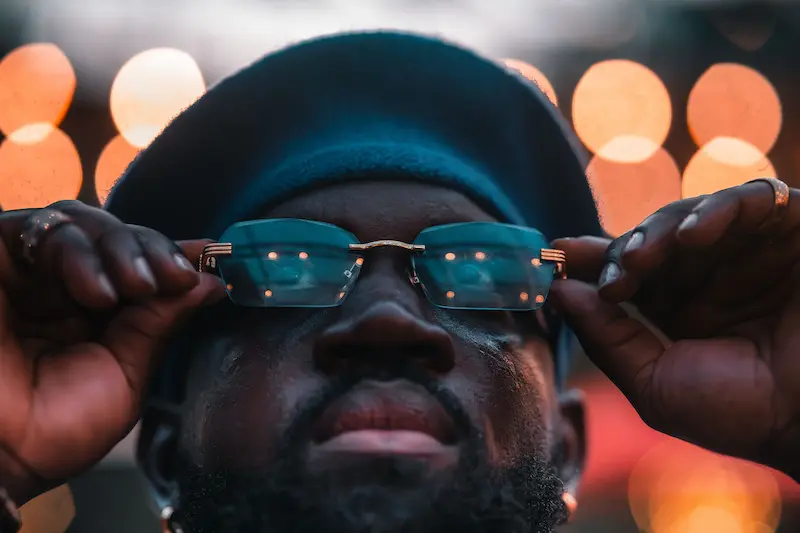 A close-up portrait of a man wearing rimless gold-frame luxury sunglasses, holding both temples with two hands to adjust them carefully