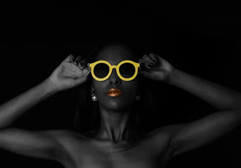 A monochrome portrait of a woman holding vibrant yellow round designer sunglasses using both hands to adjust the frame gently