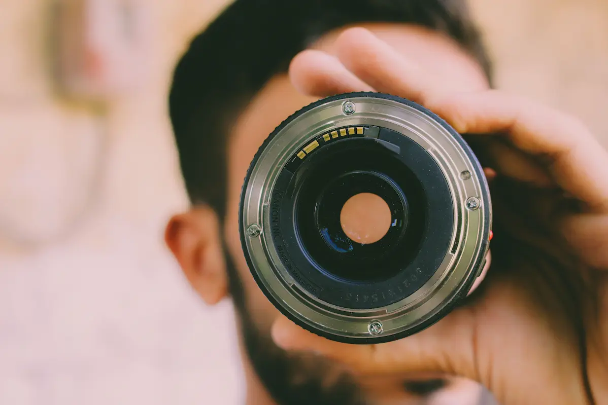 A person holding a used DSLR camera lens up to the eye to inspect the rear glass element and contacts for fungus growth