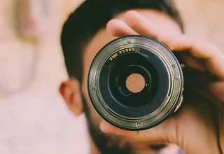 A person holding a used DSLR camera lens up to the eye to inspect the rear glass element and contacts for fungus growth