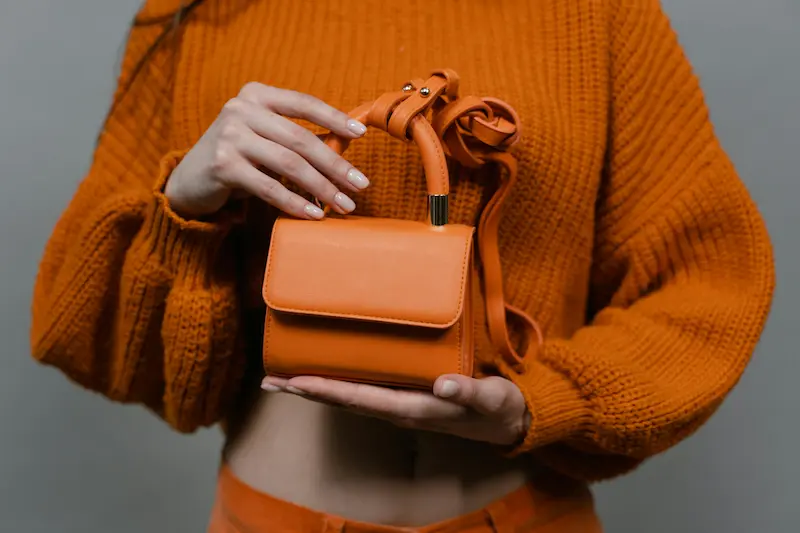 Orange leather luxury handbag with gold hardware held in hands, representing the smooth leather finish that requires species-specific conditioning protocols to maintain suppleness and prevent micro-cracking