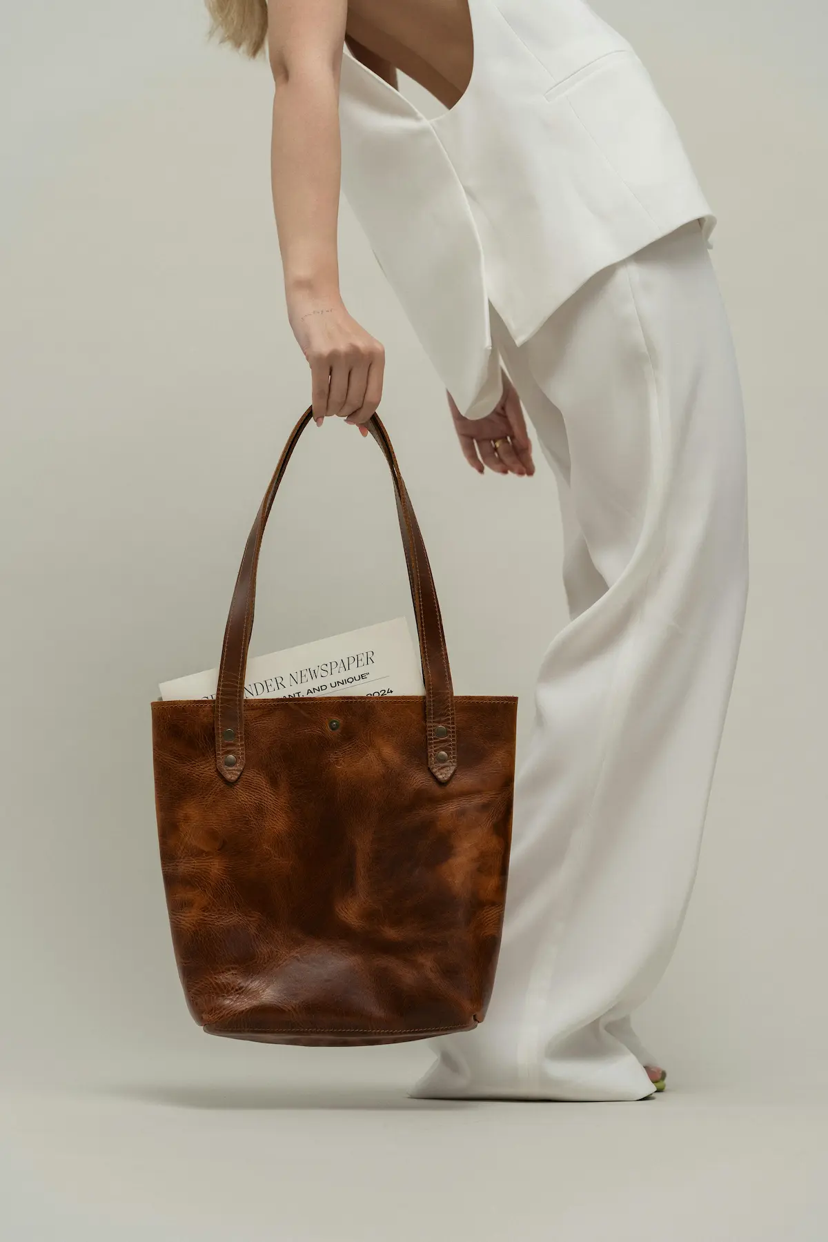 Woman in monochromatic white outfit carrying an unbranded brown leather tote bag, embodying the quiet luxury aesthetic where material quality and understated elegance replace conspicuous branding