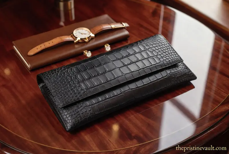 A sophisticated flat lay on a wooden table featuring an unbranded black crocodile-embossed leather clutch with matte gold hardware next to a leather watch, perfect for quiet luxury trends in 2026