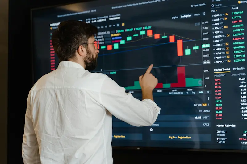 Financial analyst pointing at market trading charts displaying investment performance data, illustrating the comparative analysis between luxury handbag appreciation and traditional stock market returns